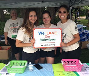 3 young women in light tan t-shirts holding a sign stating "We Love Our Volunteers"
