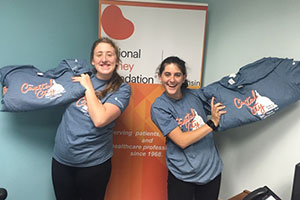 two young ladies in heather blue t-shirts each holding heather blue t-shirts with the 2019 Capital City Run/Walk logo in front of a NKFW sign