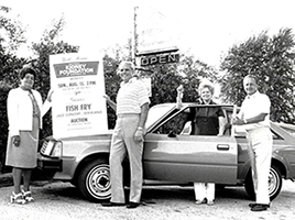 historical black and white photo of two couples standing along side a car holding a sign announcing a fish fry
